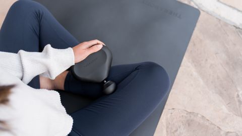 A photo of a woman using the Theragun Mini sat on a yoga mat 