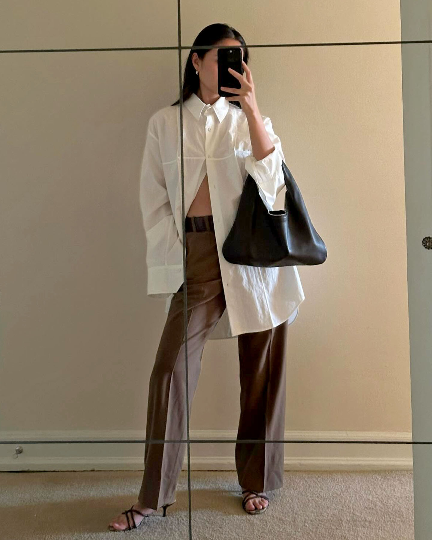 a stylish women poses for a mirror selfie wearing an oversize white button-down shirt, minimalistic black shoulder bag, belted brown trousers, and strappy black sandals