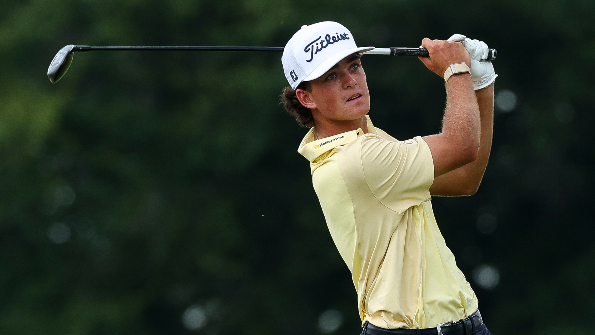 Jackson Koivun holds his finish on a fairway wood shot while wearing a yellow polo and white Titleist cap