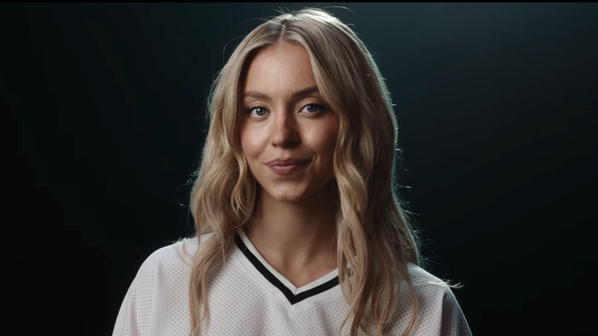 Sydney Sweeney smiling into the camera wearing a white football jersey in Novig ad