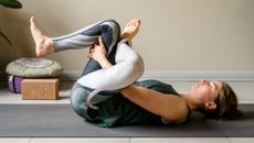 A woman practices a figure four yoga stretch at home, reclining on her back. Her eyes are closed and we see a yoga block and cushion behind her.