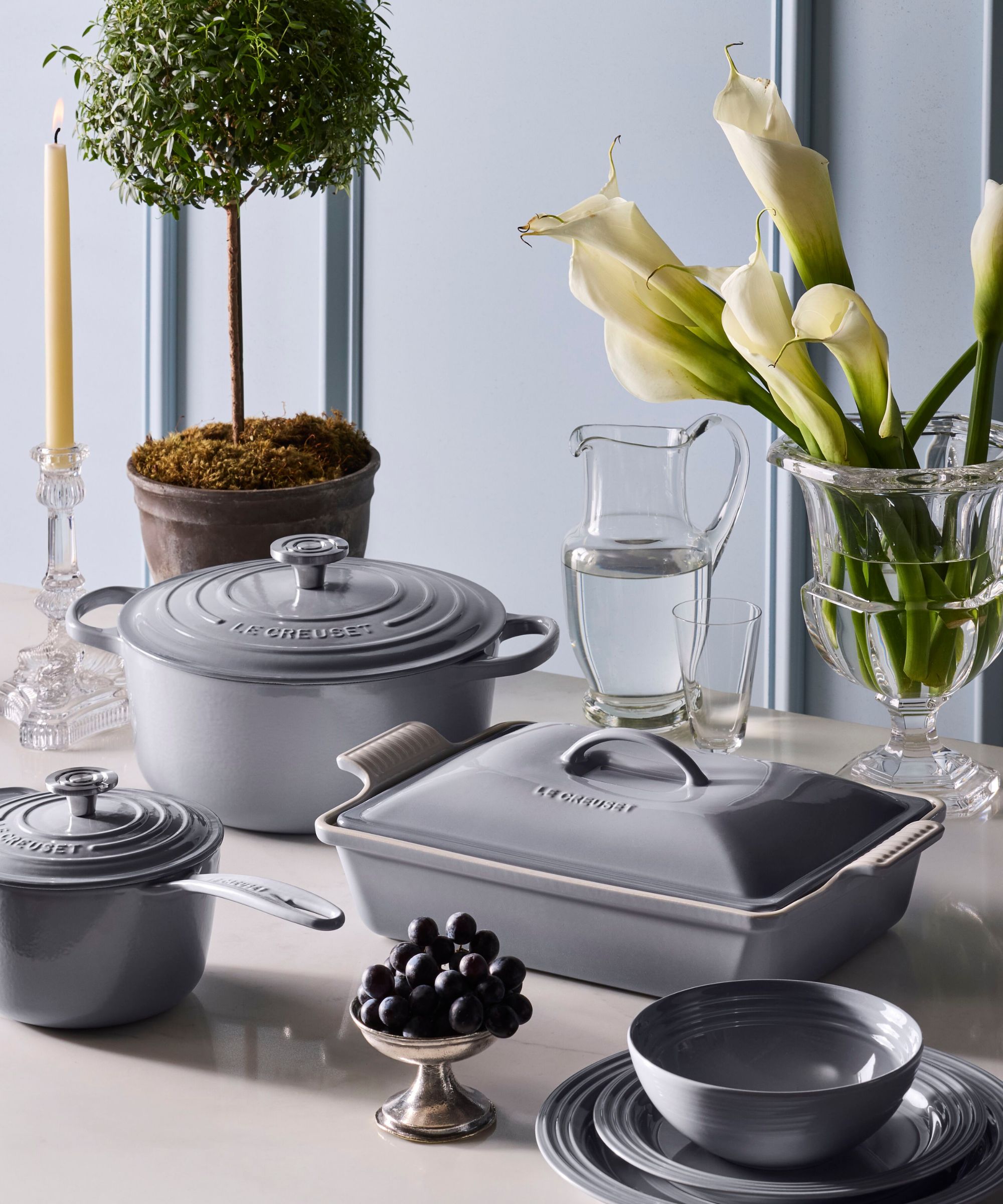 A Le Creuset Dutch oven, lidded baking dish, and small pan on a white table in a soft dove grey against a similar wall background.