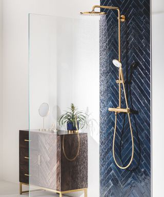 Shower stall with gold fittings and black herringbone tile in bathroom with white walls and chest of drawers