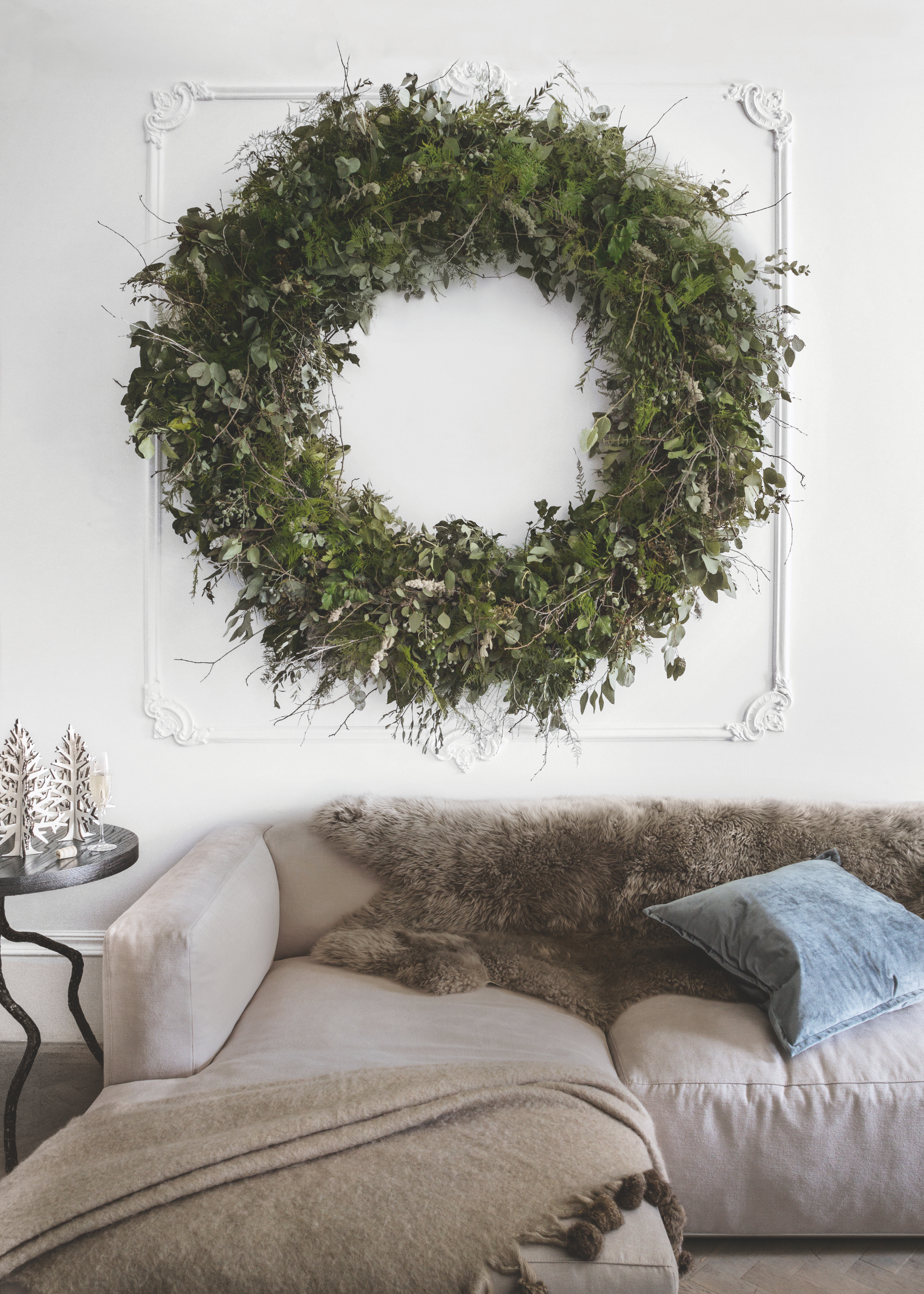 A giant natural wreath is displayed on a white wall above a beige corner sofa draped in fluffy throws with a blue pillow and a side table with tabletop christmas tree decorations