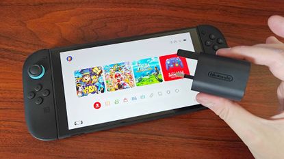 Hand holding Switch 2 power adapter next to handheld on desk.