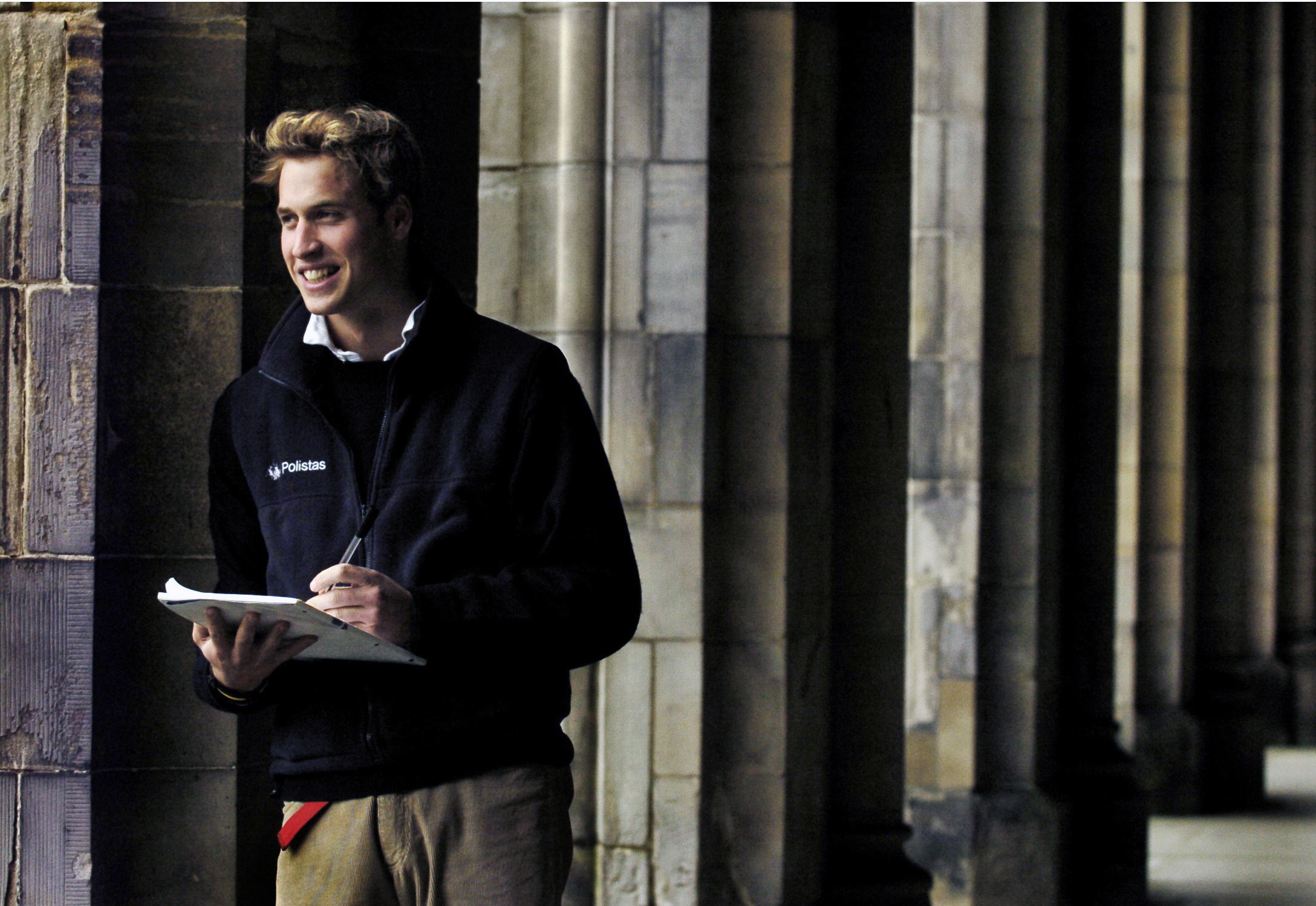 Prince William leaning on a column holding a notebook at St Andrews
