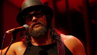 Jason Momoa wearing a black hat and sunglasses, standing in front of a microphone, in 2025