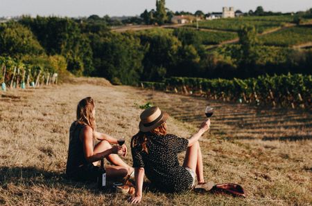 Two women sat on the edge of a vineyard with glasses of red wine
