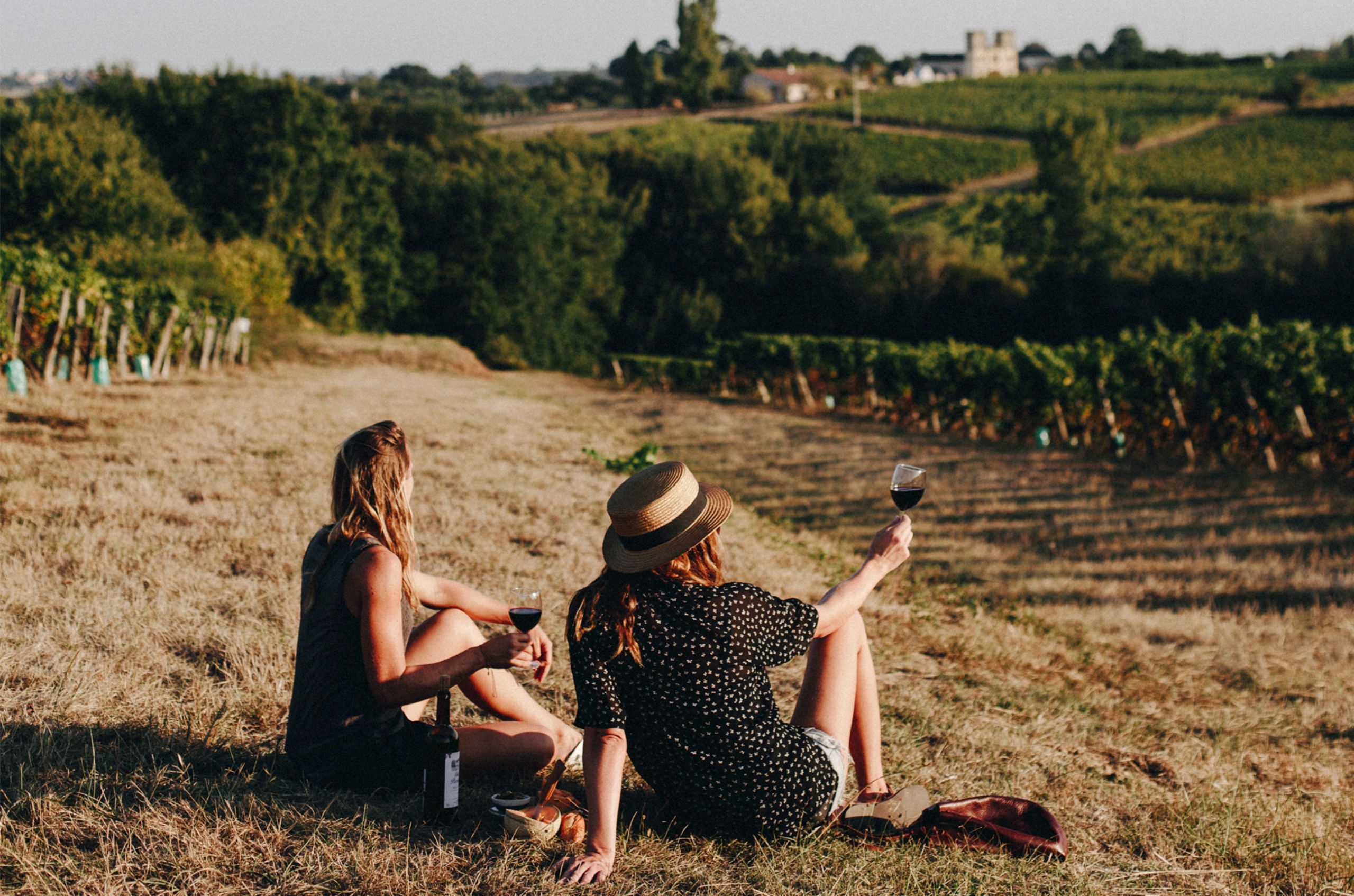Two women sat on the edge of a vineyard with glasses of red wine