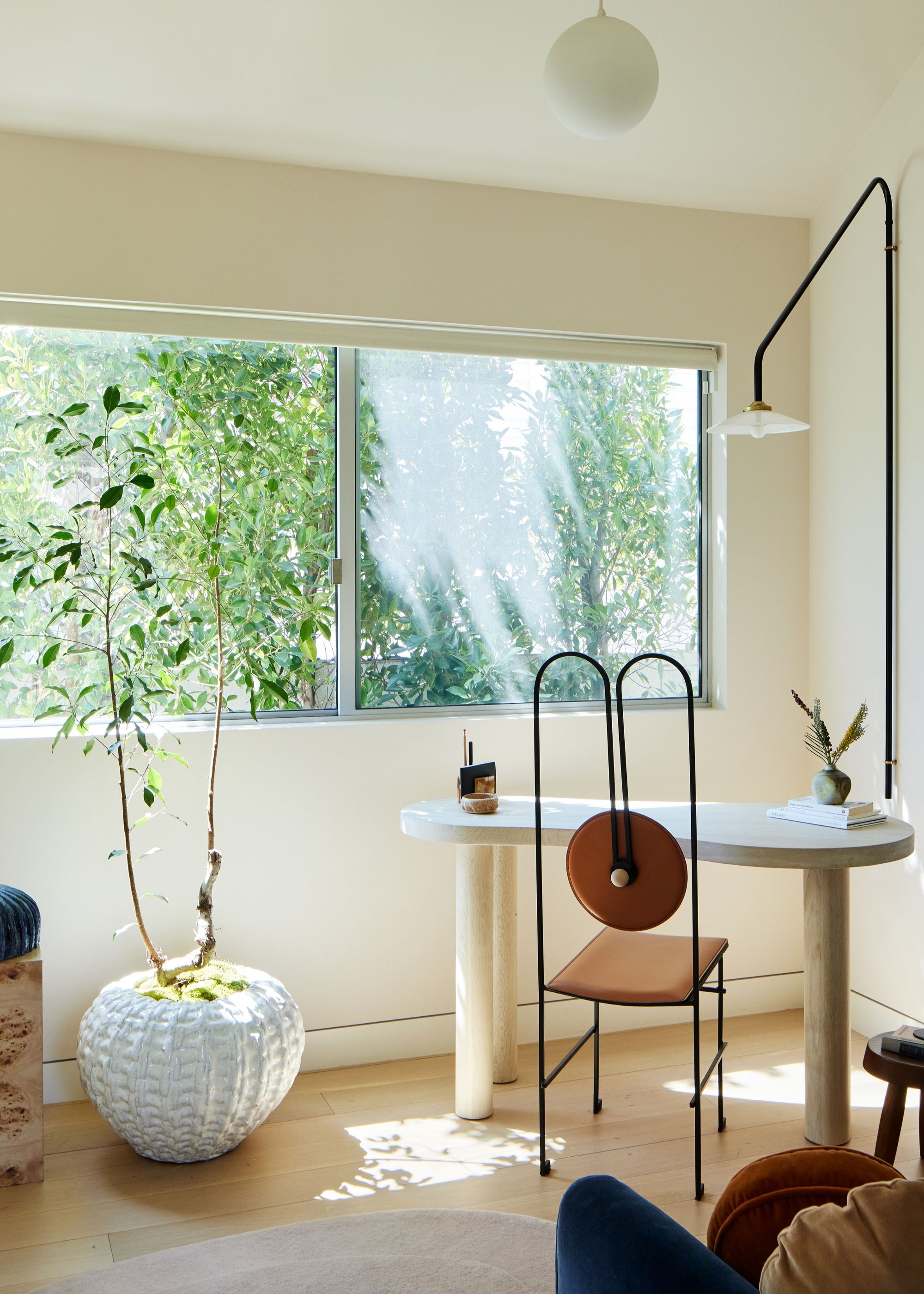 A white study with large windows, a potted tree, a small deks, a metal chair, a wall lamp, and a vase of stems