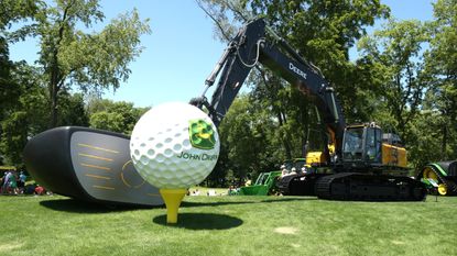 John Deere excavator with a oversized clubhead where the grabber goes, with this head lined up behind an oversized ball on top of a tee 