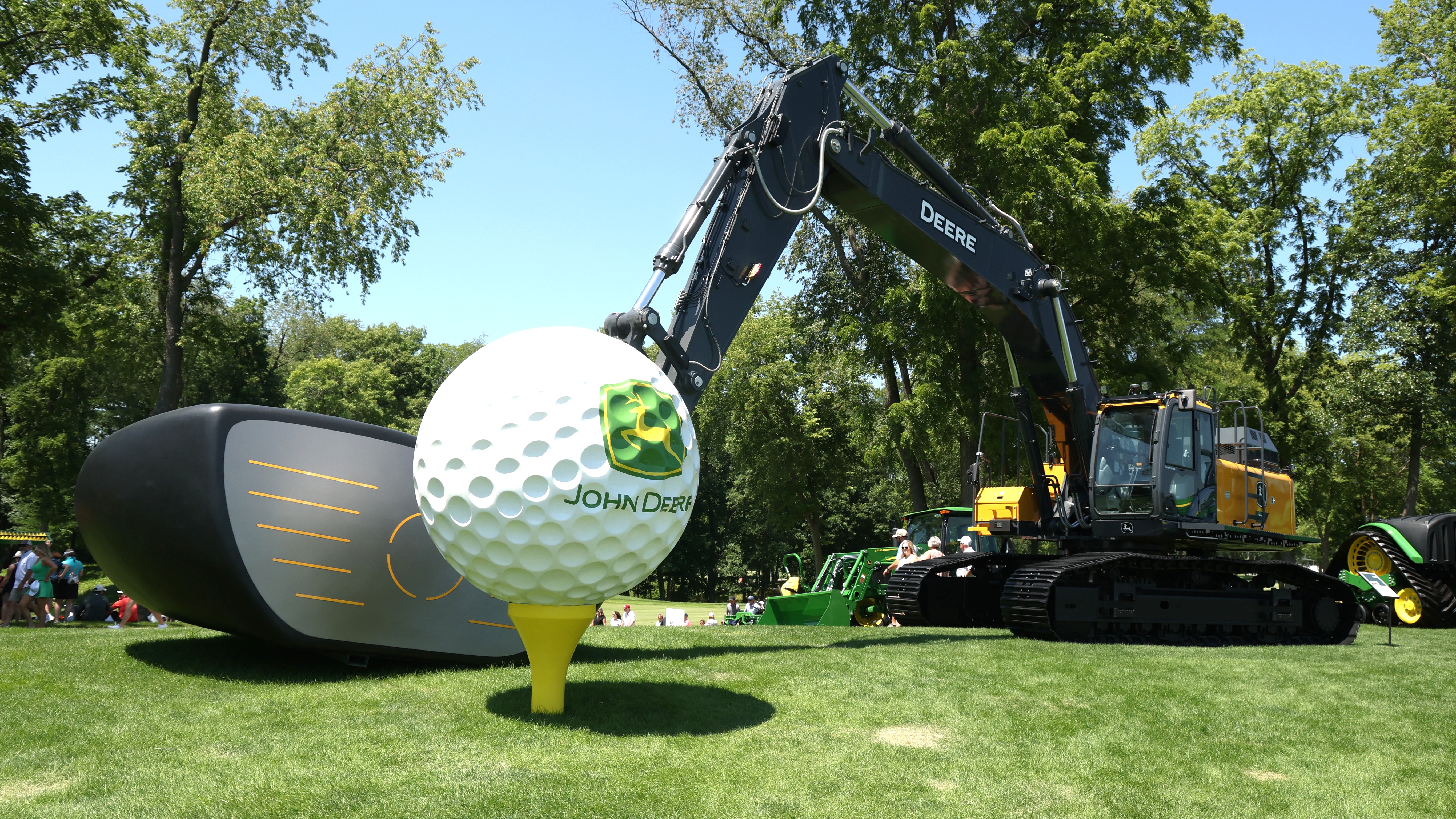 John Deere excavator with a oversized clubhead where the grabber goes,  with this head lined up behind an oversized ball on top of a tee 