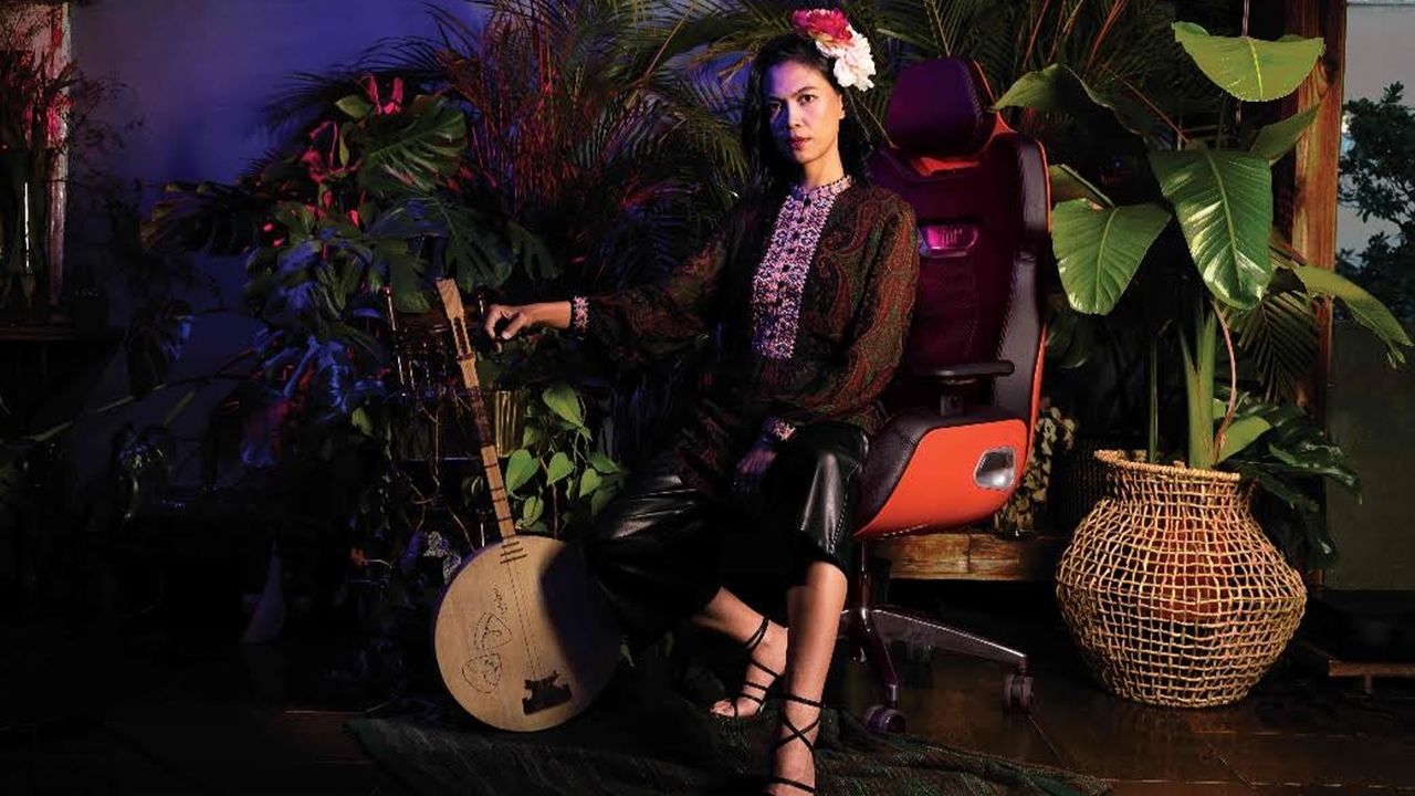 A lady sits on an orange chair amongst hordes of plants, holding a mandolin.