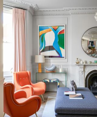 Gray painted living room with paneling, framed colorful painting, pink curtains and twin orange lounge chairs, glass console table and marble fireplace, large blue ottoman,
