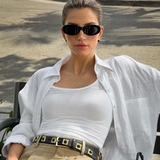 female fashion influencer Cass Dimicco sits on a bench outside wearing black oval sunglasses, white linen button-down shirt, white tank top, embellished belt, and tan trousers
