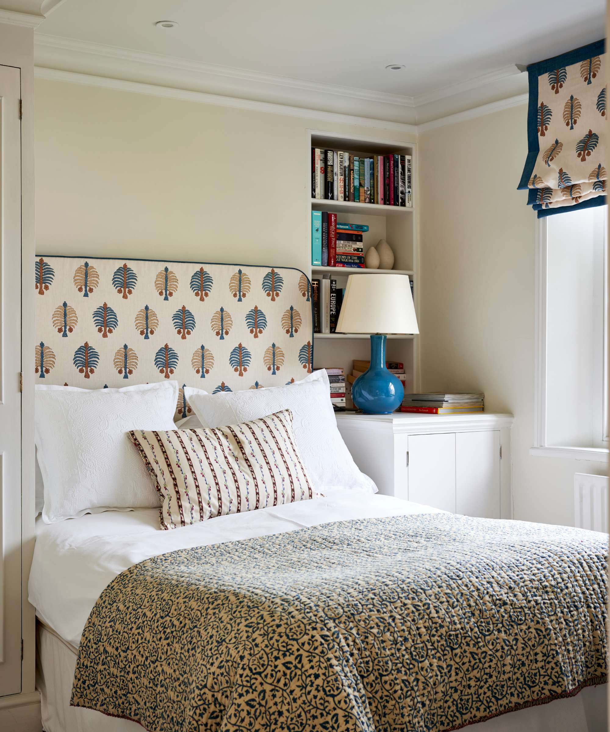 a bedroom with a quilt on the bed, a patterned headboard, built-in bookshelves on the wall on the right, and a blue lamp