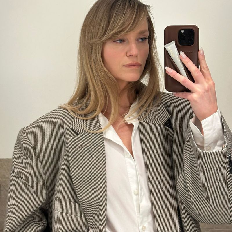 A woman with a bouncy blowdry and fringe taking a mirror selfie with a Rhode phone case