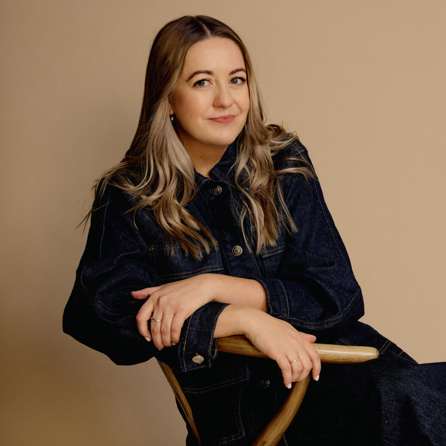 A headshot of a woman with long brown-blonde hair, sitting in a wood chair