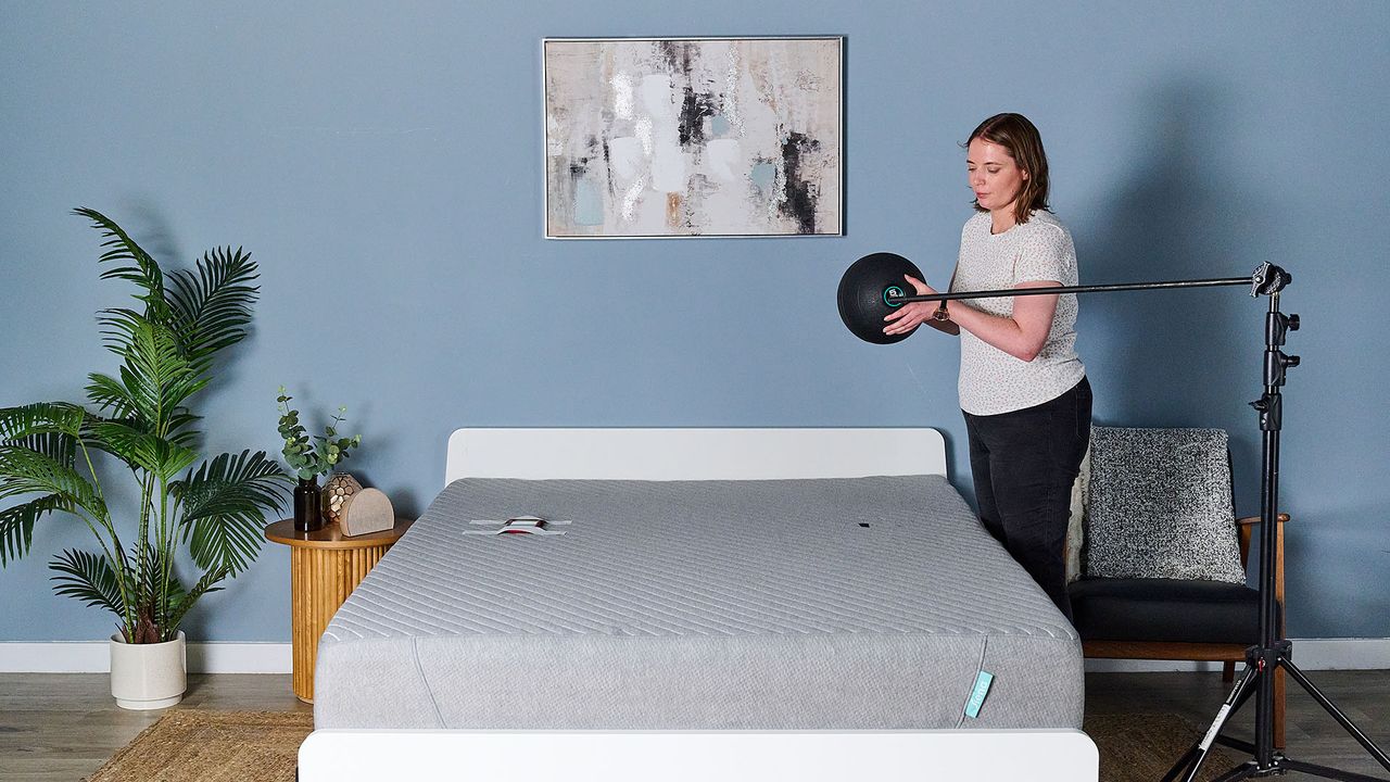 This image shows the preparation for our objective slam ball motion isolation test in our sleep studio. The Siena Memory Foam Mattress on a bed frame sits in the center of a decorated room. To the right of the bed, our sleep writer Ruth Jones holds a 5kg slam ball over the top of the bed. Next to her, a rig is set to mark 24 inches from the surface of the bed. On the left of the mattress is a mark roughly where a body would be when sleeping &mdash; this is the drop zone. On the left of the mattress, in the equivalent sleeping area, a phone with an accelerometer app open is taped to the bed.