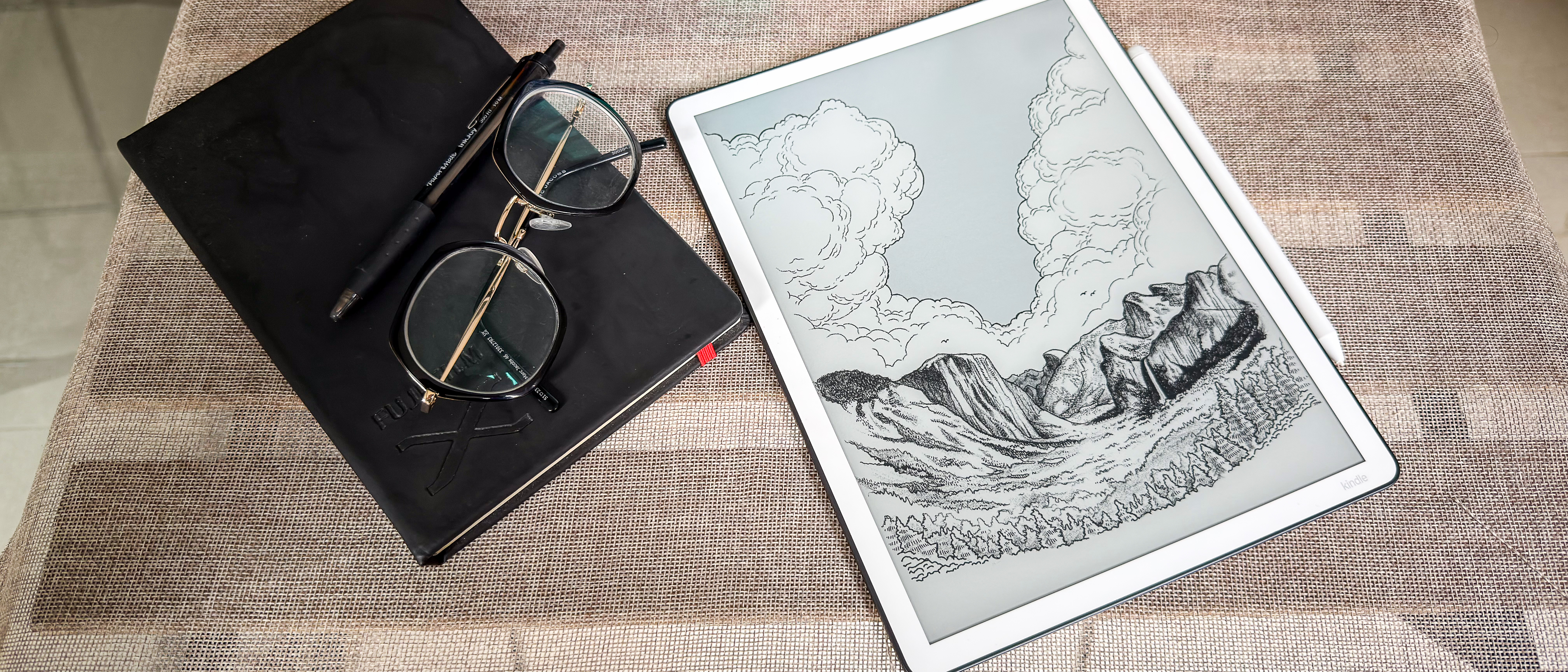 The 3rd generation Kindle Scribe showing a wallpaper on its screen lying on a table beside a notebook, spectacles and pen
