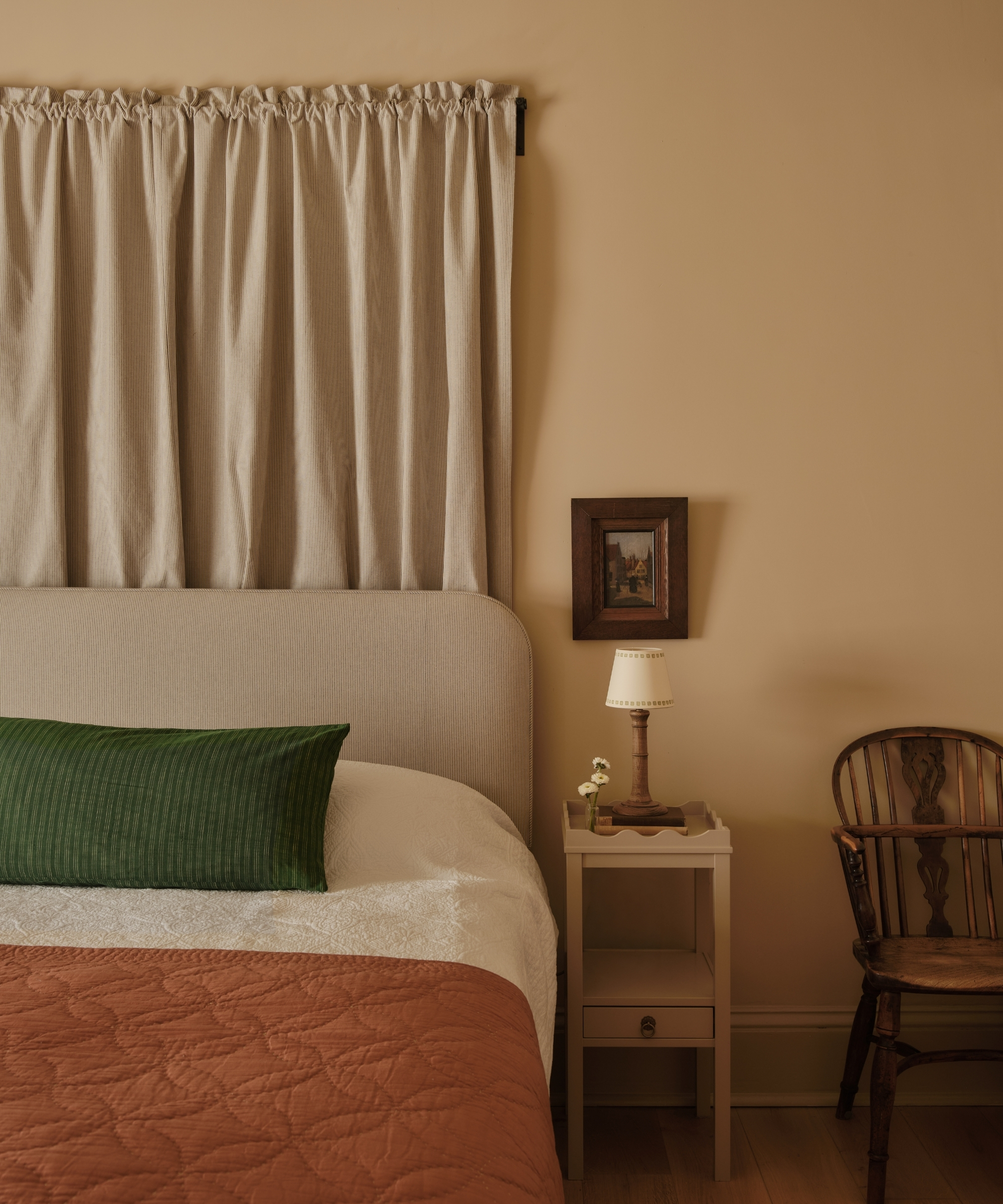 Warm bedroom with layered white bedding, rust quilt, green pillow, linen curtains and antique wooden chair.