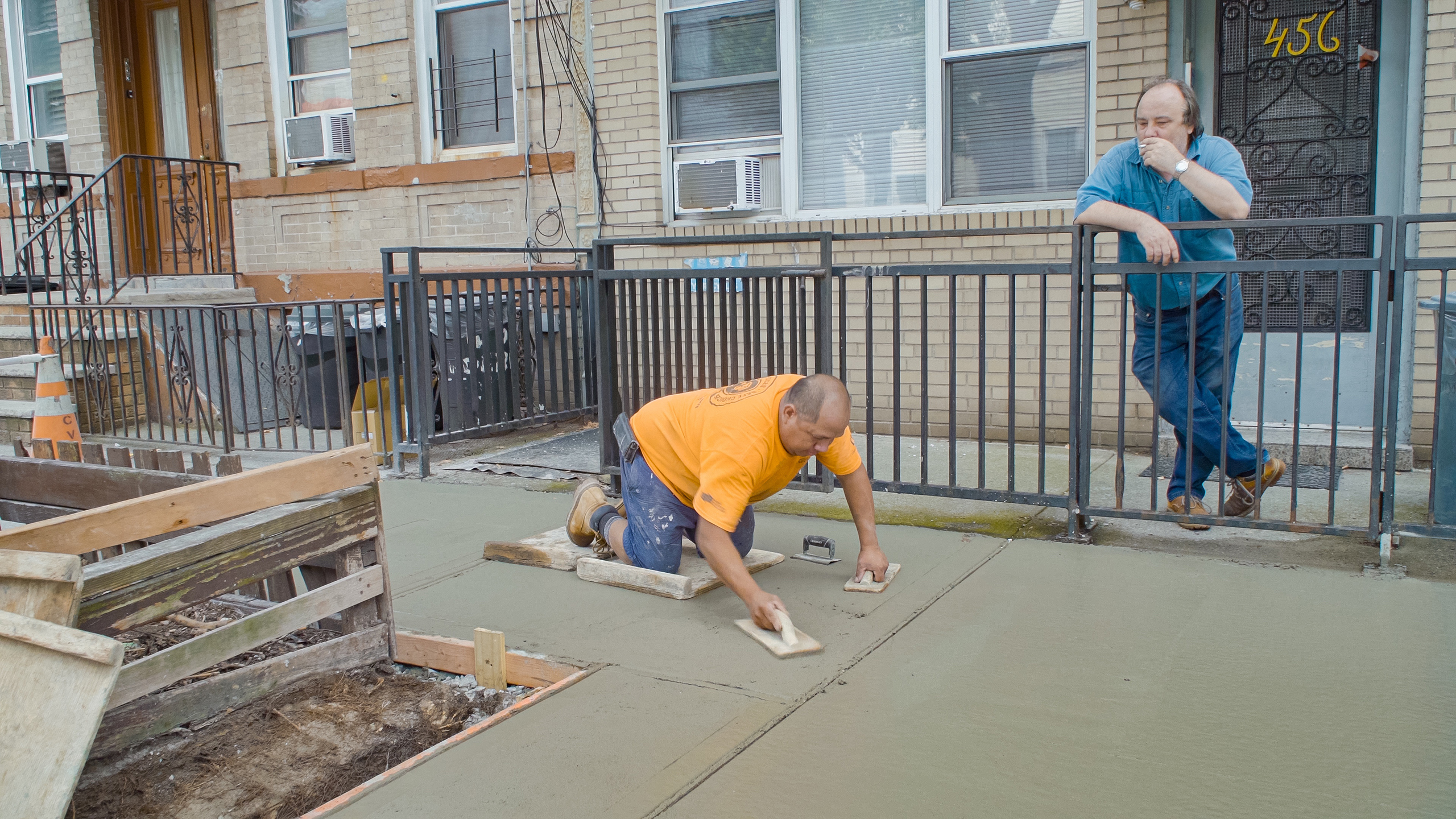 a man in a yellow shirt lays down concrete while another man watches in front of an apartment front door in a still from the john wilson documentary history of concrete