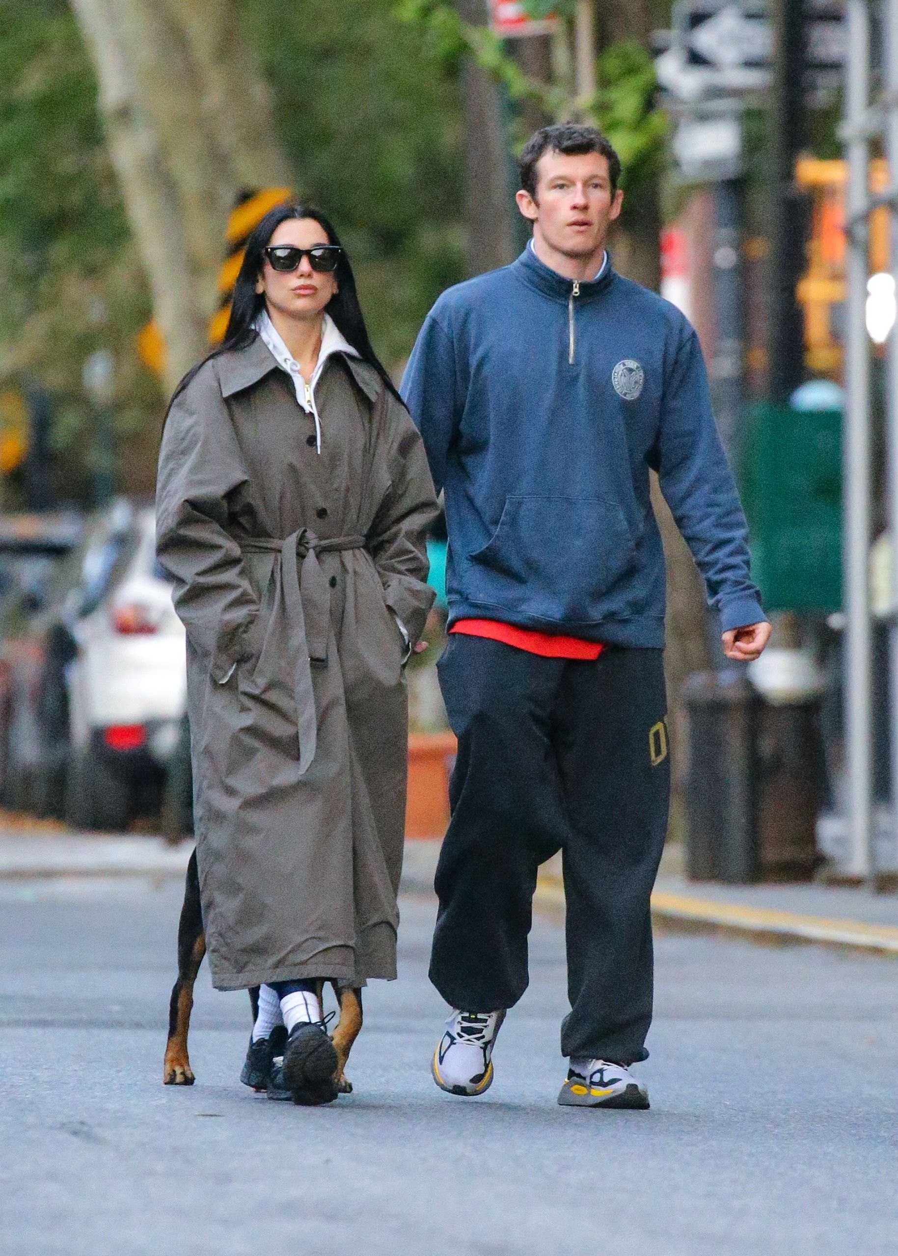 Dua Lipa walks down the street with Calum Turner wearing a grey single-breasted trench coat with leggings, a hoodie and black trainers.