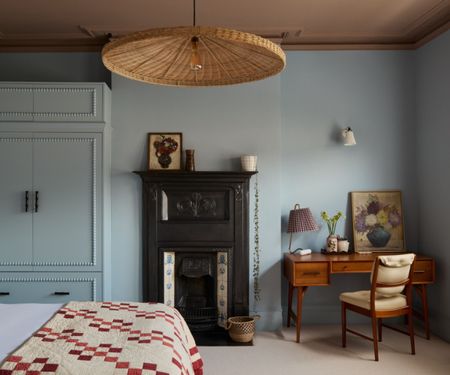 Bedroom with pale blue walls and plaster pink ceiling, large rattan pendant light, fireplace, and midcentury desk in the corner with artwork and pleated table lamp. A bed features a vintage red and cream quilt.