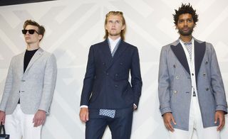 Three guys wearing the Hardy Amies S/S 2015 collection. On the left the guy is wearing a navy t-shirt, gray jacket, white pants and sunglasses. Next to him the guy is wearing a white shirt, navy suit with a blue scarf and sunglasses on his head. and on the right the guy is wearing a gray shirt, white jacket, white pants and a navy overcoat
