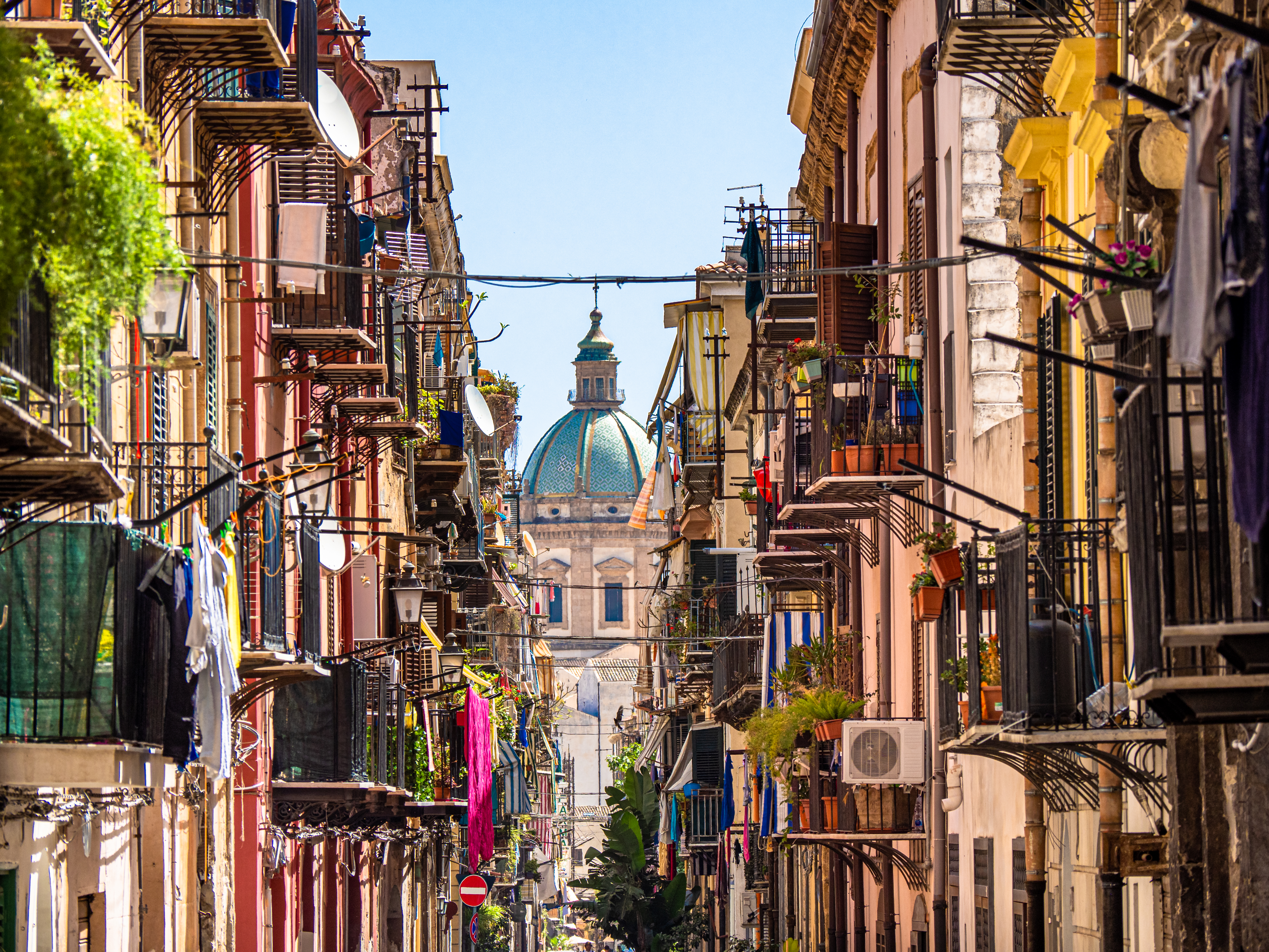 Narrow street of high houses with balconies strewn with washing and the dome of a cathedral in the background