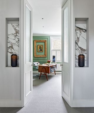 Open door flagged by rectangular marble alcoves looking through to study with retro swivel chair and desk with green painted feature wall, gray carpet