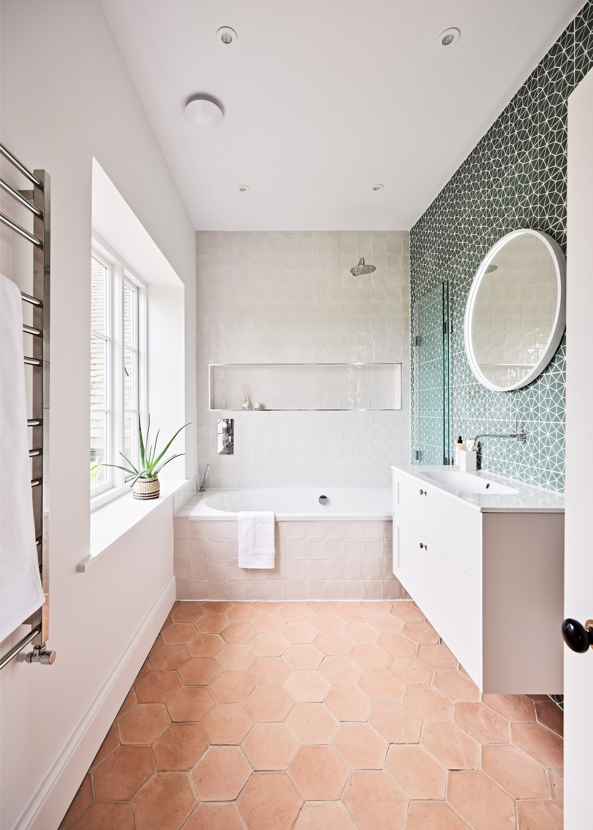 A small, predominantly white bathroom with a built-in tub, hexagon terracotta floor tiles, a green patterned tile accent wall and floating vanity unit