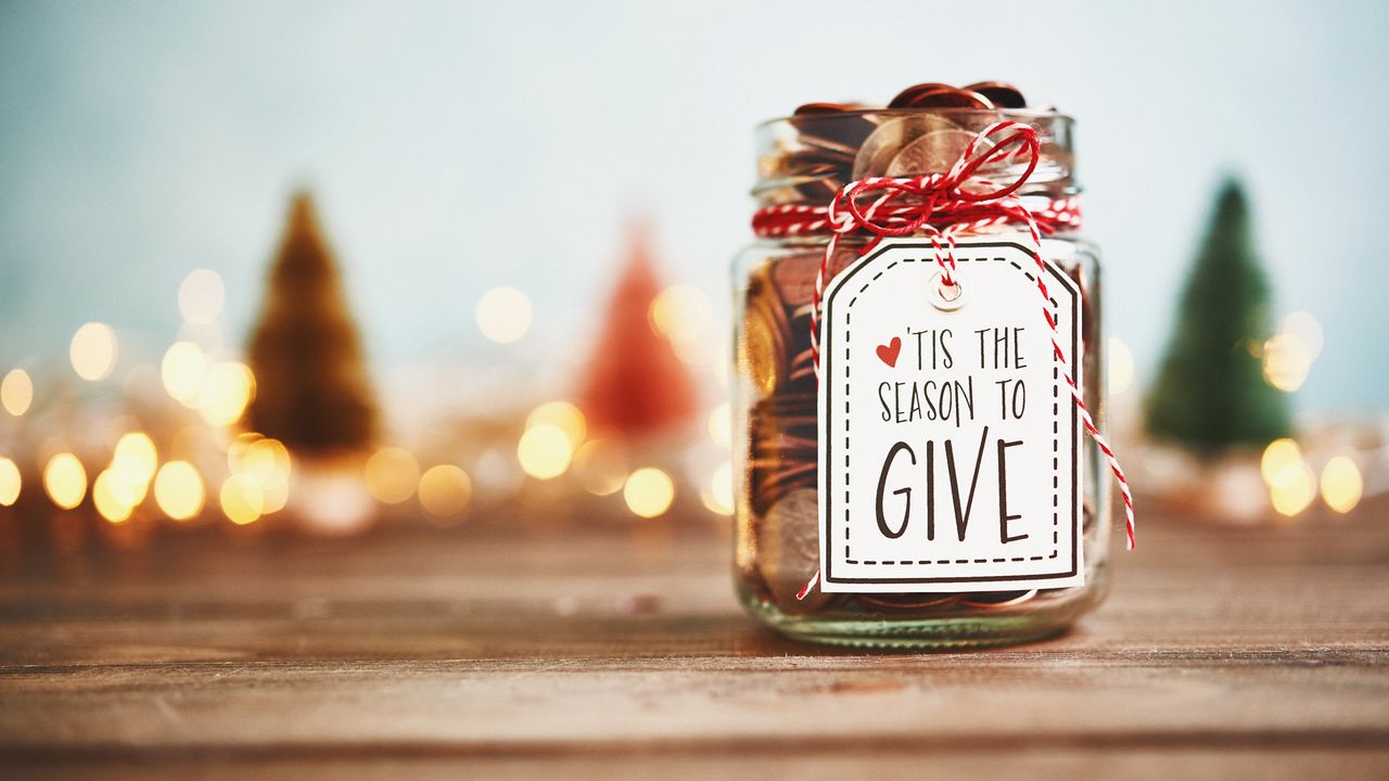 A glass jar labeled &#039;Tis the Season to Give sits on a table with holiday decor in the background.