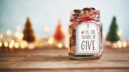 A glass jar labeled 'Tis the Season to Give sits on a table with holiday decor in the background.