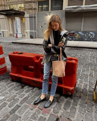 Courtney Grow wears jeans, black loafers and leather jacket, grey sweater and The Row bucket bag in New York.