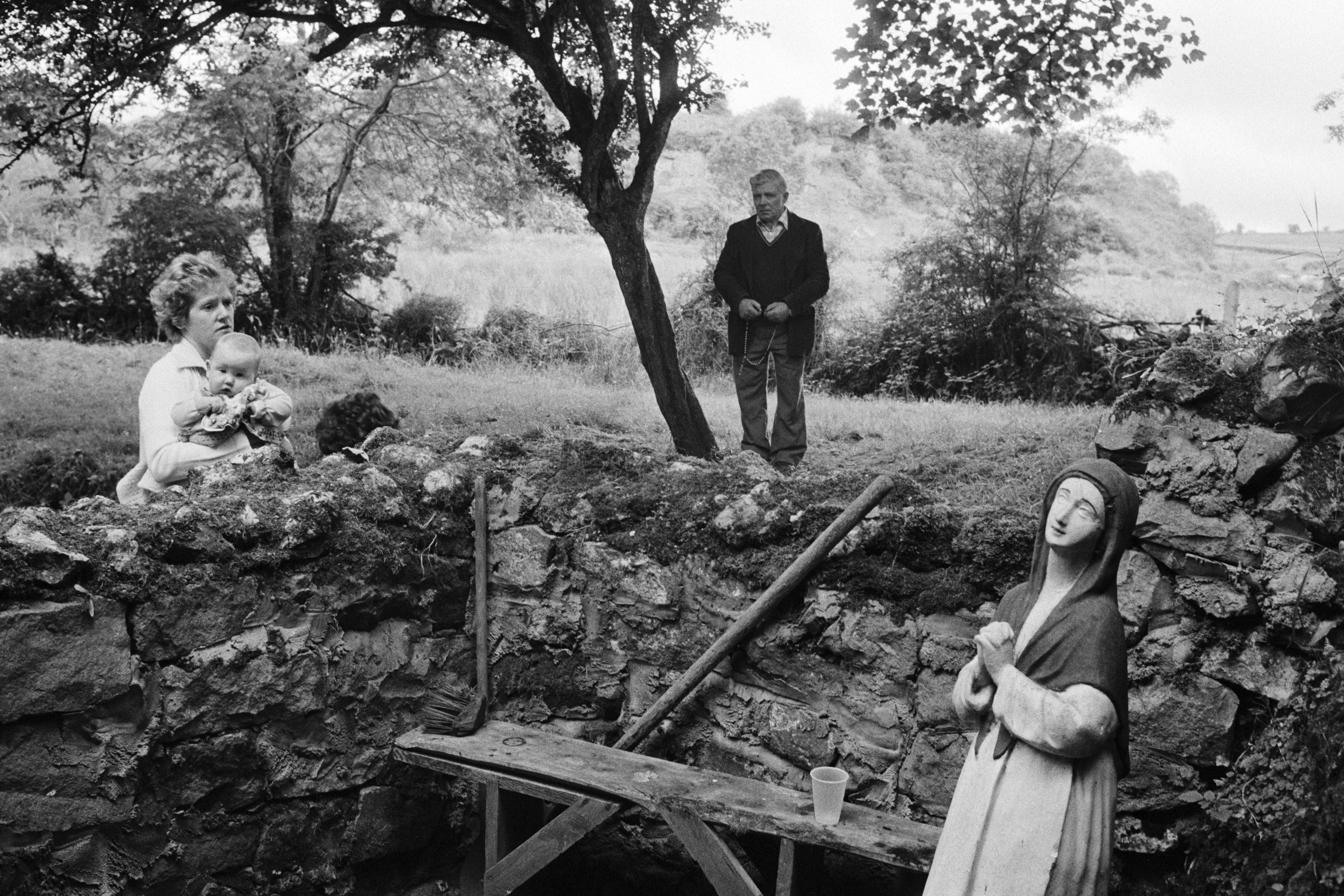 A woman holding a baby and a man standing in a field look toward a religious statue placed within a rustic, stone-walled enclosure.
