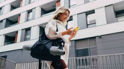 A woman in a city is walking and looking at her phone. She wears an oversized jumper and cycling shorts and has a gym bag slung over one shoulder, with a yoga mat in it. 