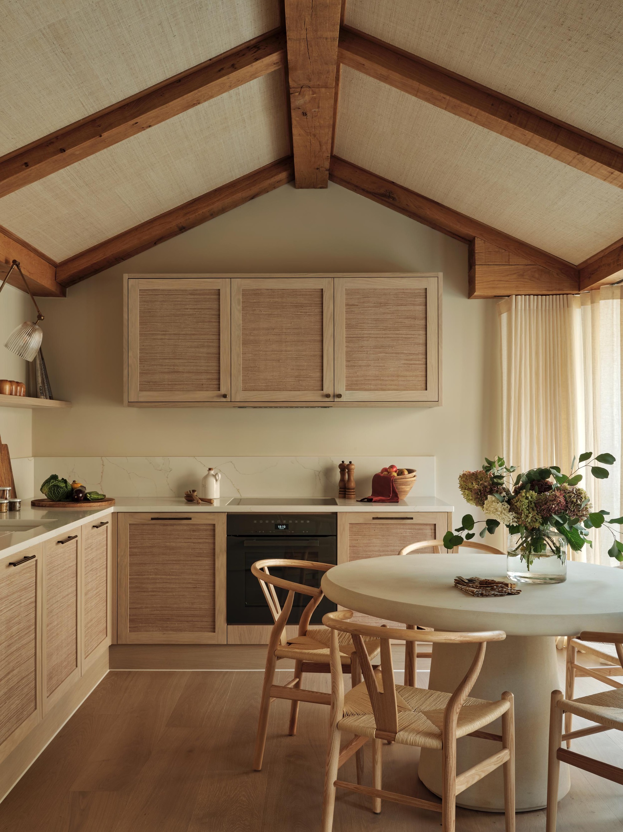 neutral kitchen-diner with white concrete round table in the middle with wishbone chairs, L-shaped kitchen with raffia fronts, timber cabinetry, and white countertops, beige walls, and a ceiling with exposed beams and raffia panels