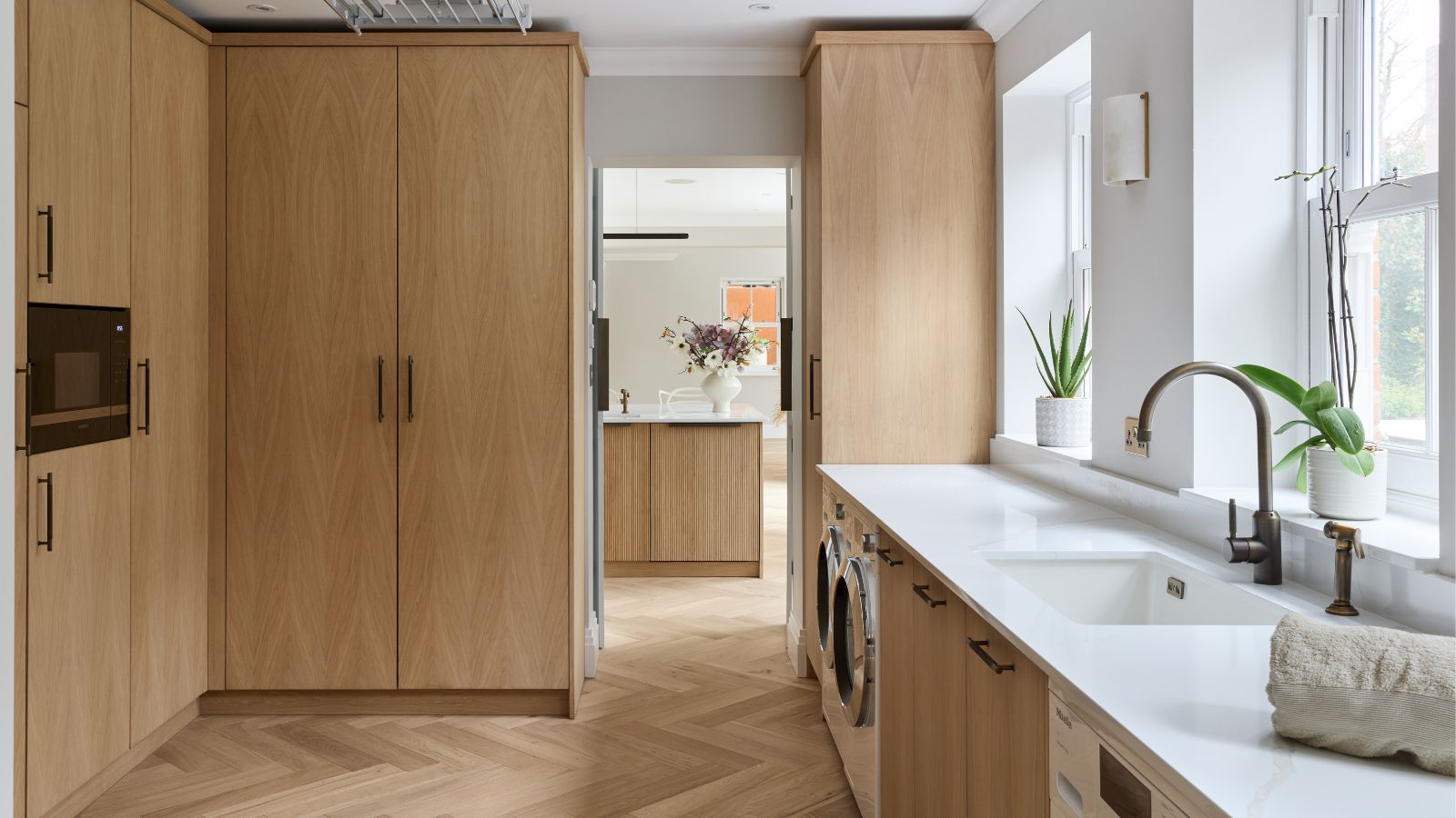 A light oak utility room with full-height cabinetry, white stone worktop, integrated washing machines, a bronze tap and oak herringbone floor tiles leading through to an open-plan kitchen.