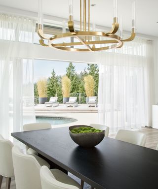 An open-plan dining are with black dining table, gold contemporary chandelier and view of a pool through floor-to-ceiling windows
