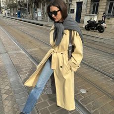 French fashion influencer Anne-Laure Mais in Paris wearing a chic fall outfit styles with blue jeans, a classic trench, sunglasses, a grey knit tied at her shoulders, and black boots.