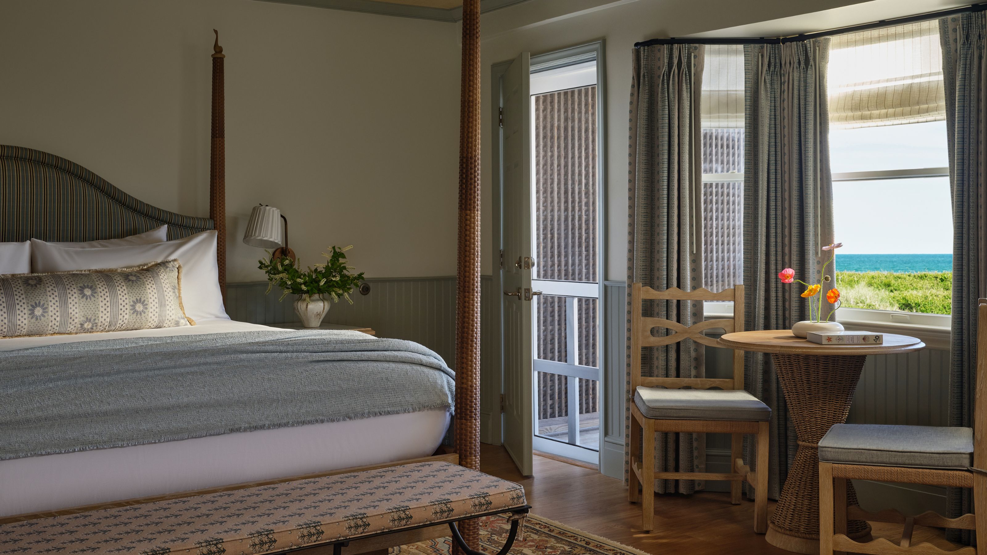 A cozy bedroom featuring a four-poster bed with a light blue blanket, alongside a small wicker table and two wooden chairs overlooking the sea.