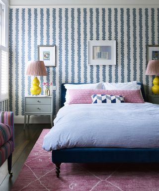 A bedroom with blue striped wallpaper, pink bedding and rug, and yellow bedside lamps