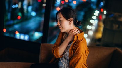 Woman sitting on sofa holds her neck with one hand. Her eyes are closed. She sits in front of a window. It is night time.