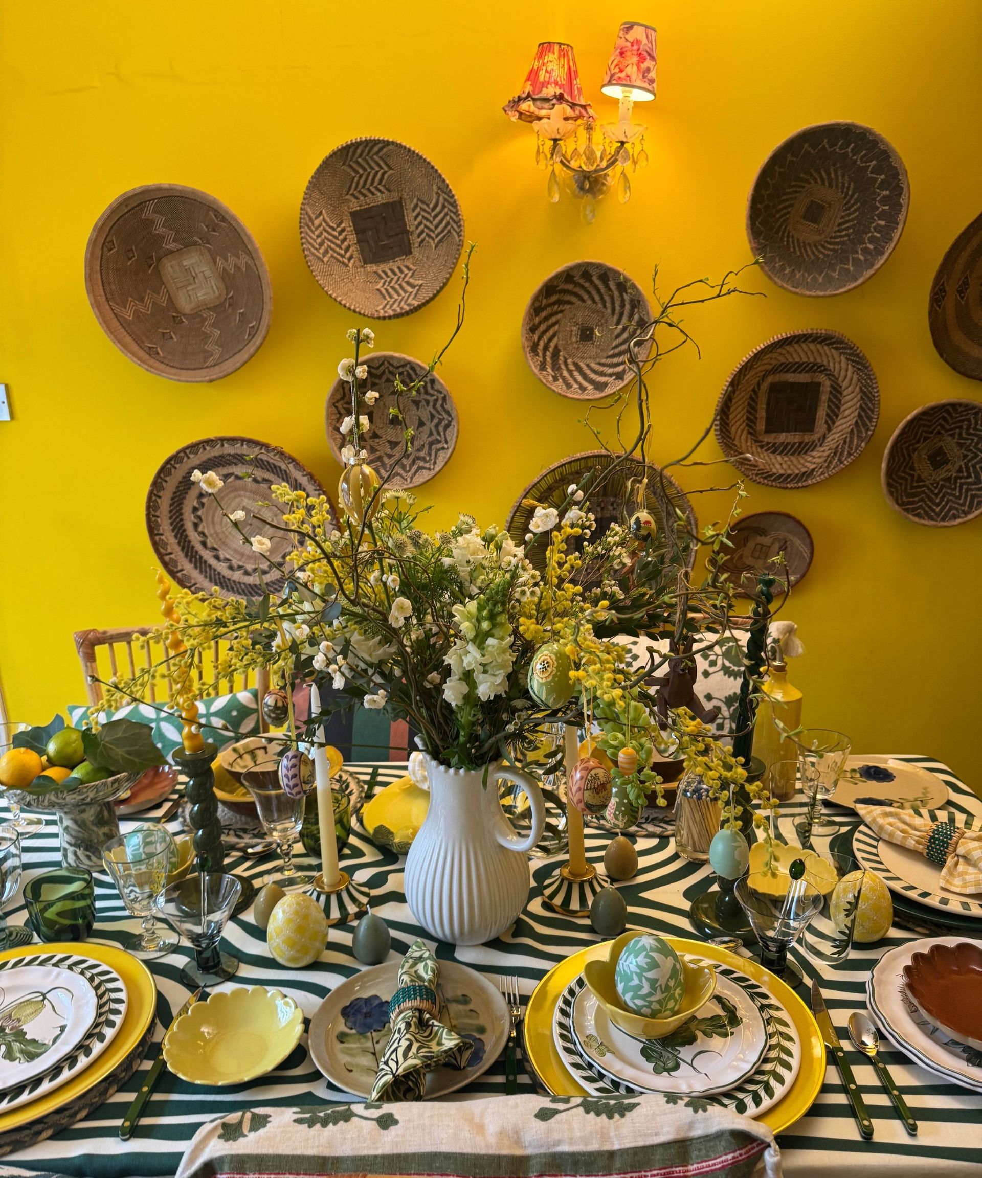 Vibrant table setting with yellow and green theme, mis-matched plates, Easter egg decorations and an Easter tree in the centre, against a yellow wall with baskets displayed on it.