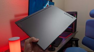 A Lenovo Legion 5i (Gen 9) gaming laptop held in the hand while closed, showing the "Lenovo" and "Legion" logos on the lid with a red backlight.