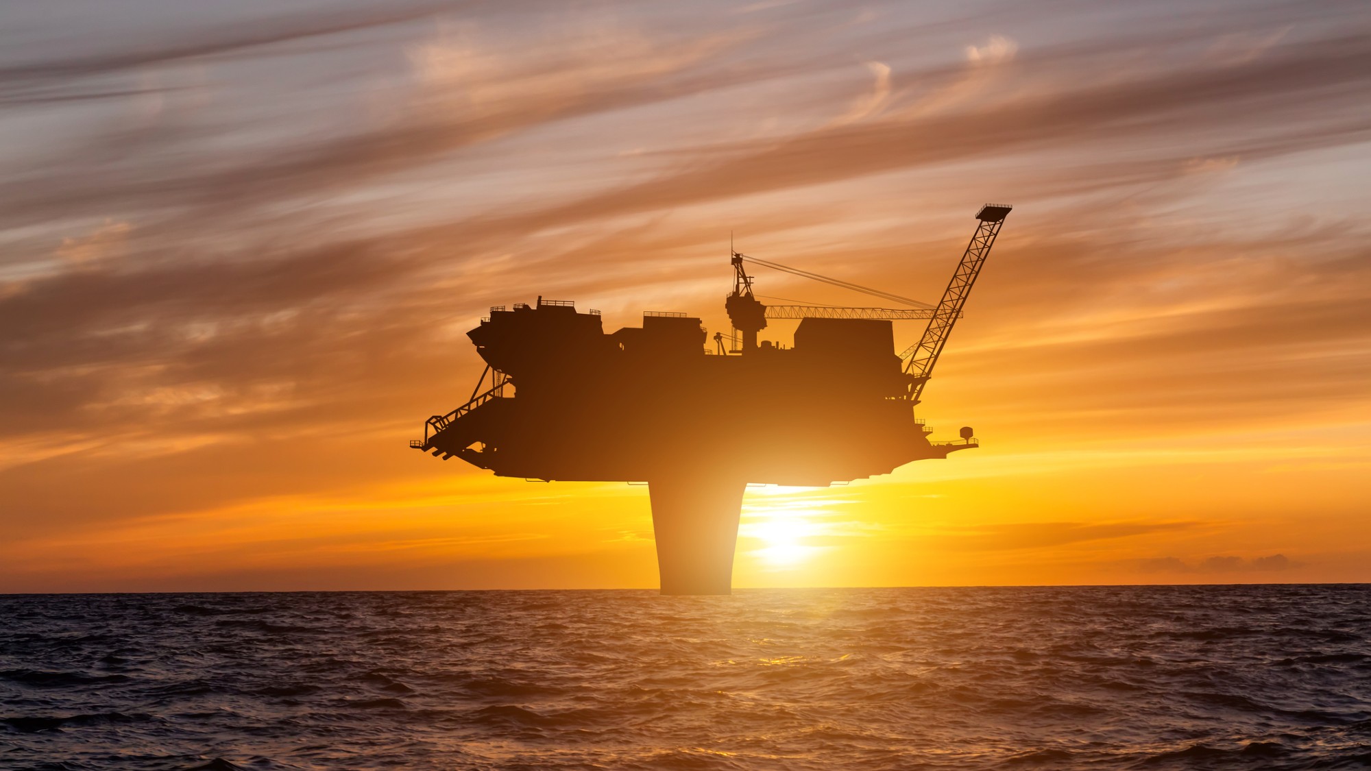 the sun sets behind a large T-shaped drilling platform at sea