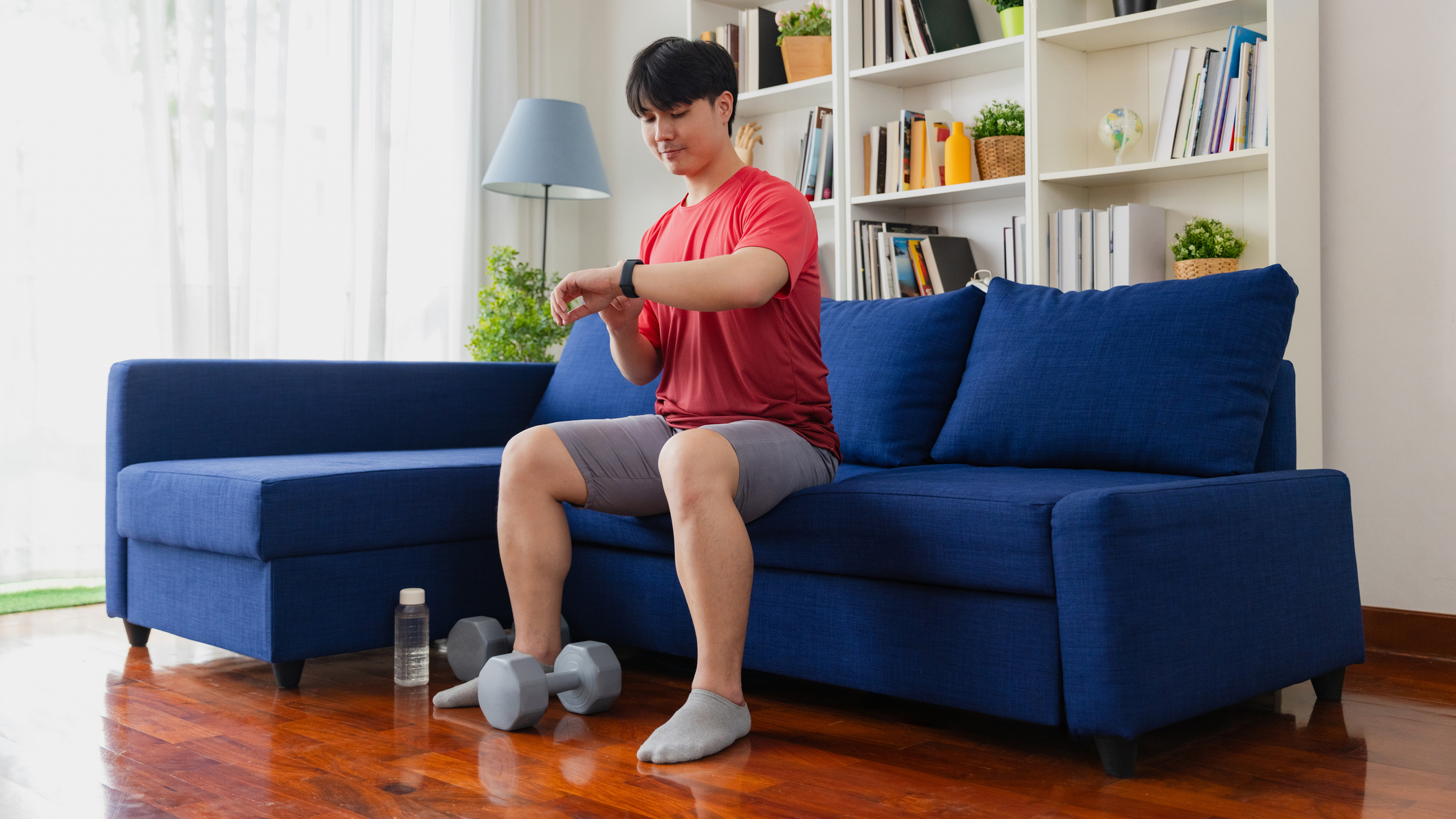 Man sitting on couch with dumbbells on the floor between his feet, he is looking at the fitness tracker on his wrist