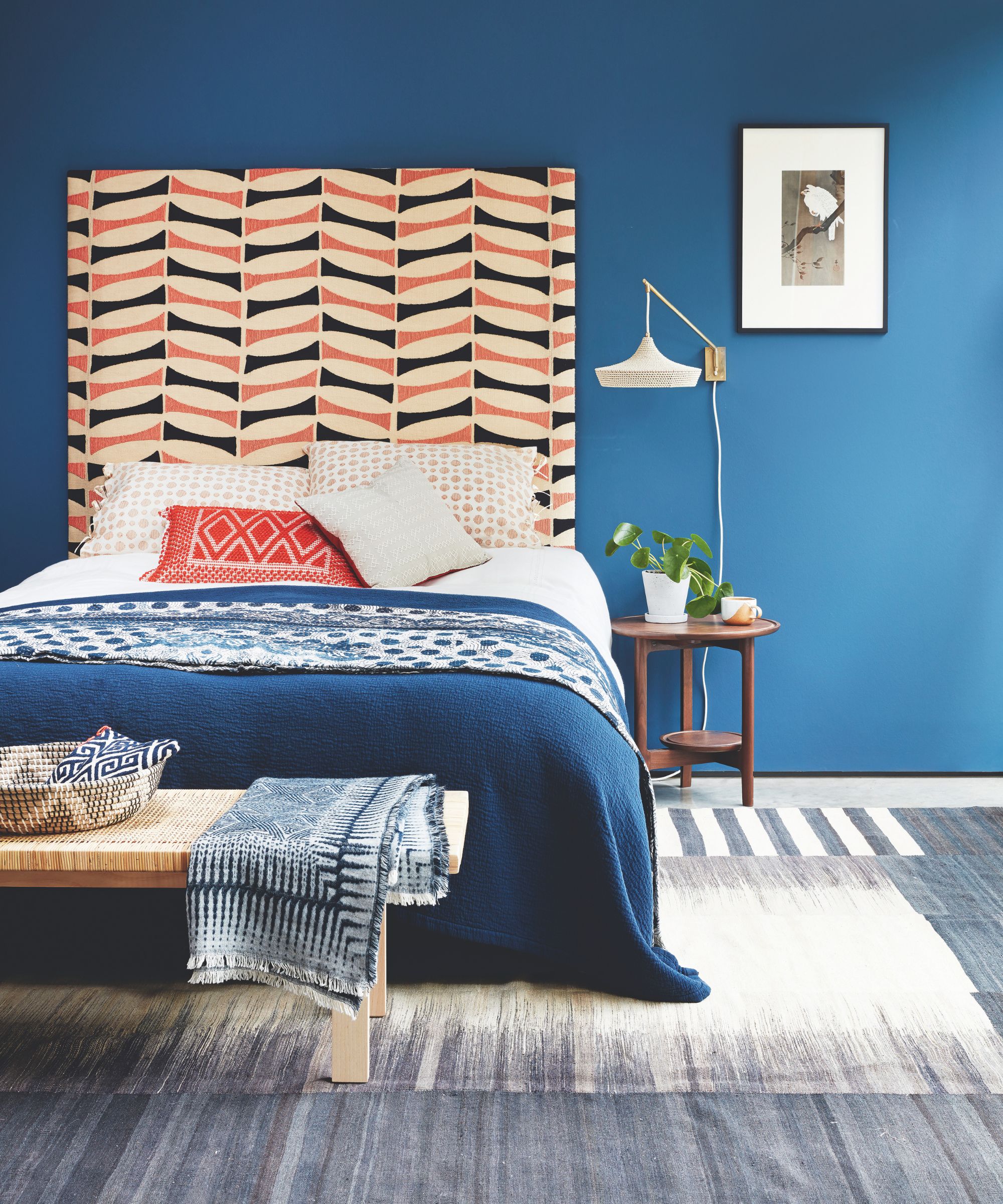 A modern bedroom with a blue wall, large statement geometric headboard, grey rug and blue bedding. Beside the bed is a wooden bedside table with a houseplant on it, and above is a small artwork in a black frame. In front of the bed is a wooden ottoman with a geometric bowl and blanket on top.