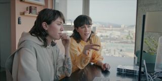 Tegan Quin and Sara Quin look at a flat desktop computer while sitting in an office in front of a floor-to-ceiling window, in 'Fanatical: The Catfishing of Tegan and Sara.'